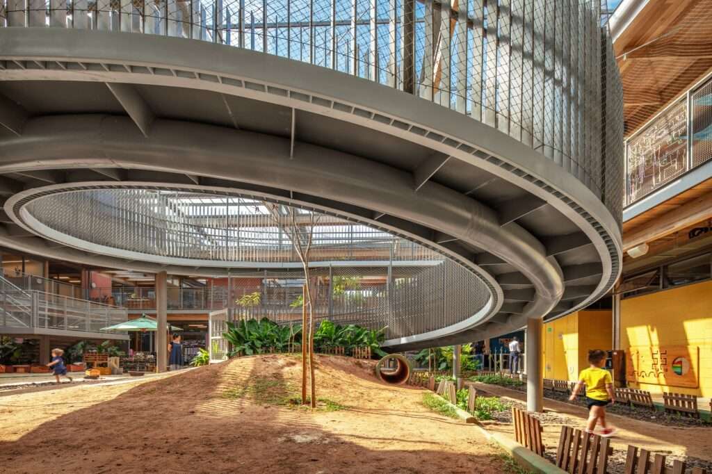 Underside view of a curved steel circular ramp in a sandy courtyard with children playing, featuring security mesh railings and timber columns.