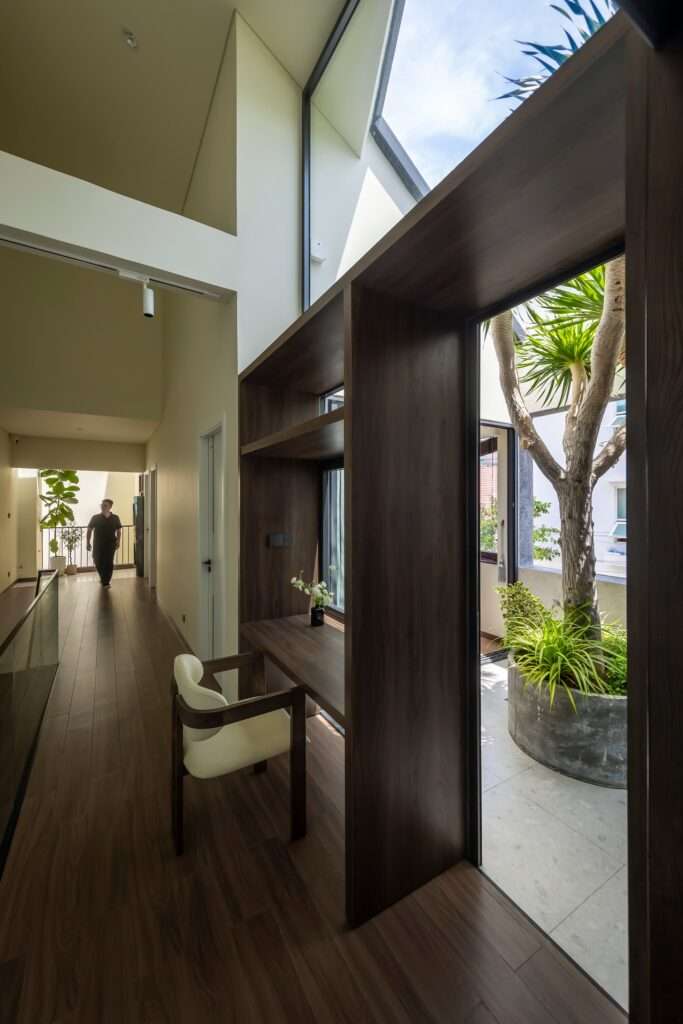 Upper-floor interior hallway and built-in wooden study desk opening directly onto a light-filled inner courtyard planter with a mature tree.