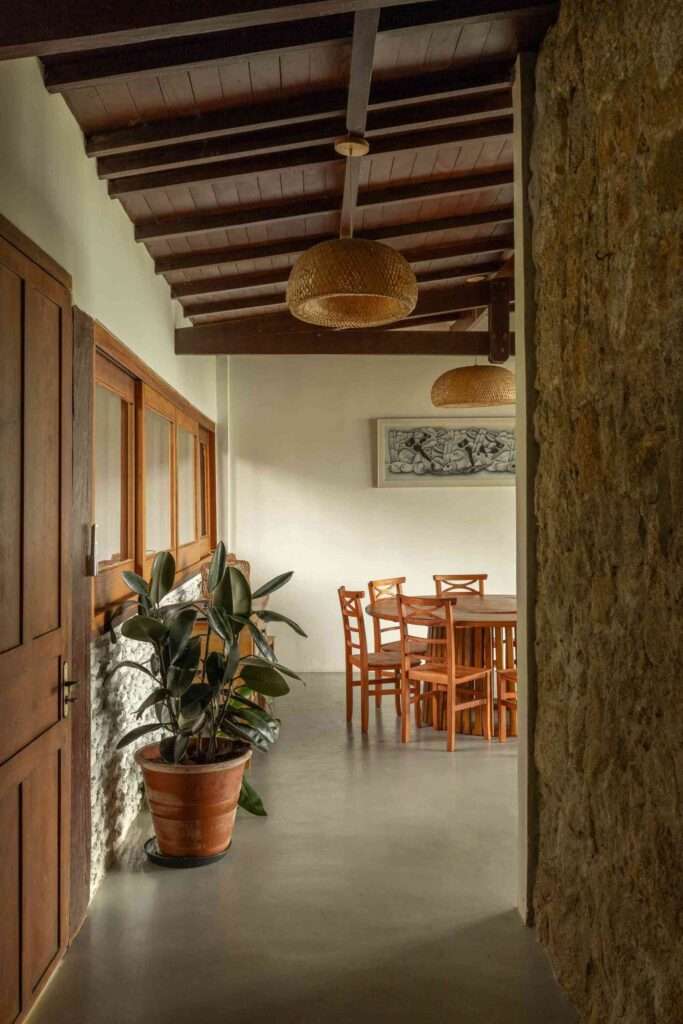 Interior dining space showing a round wooden table, rattan pendant lamps, smooth concrete flooring, and an original stone accent wall.