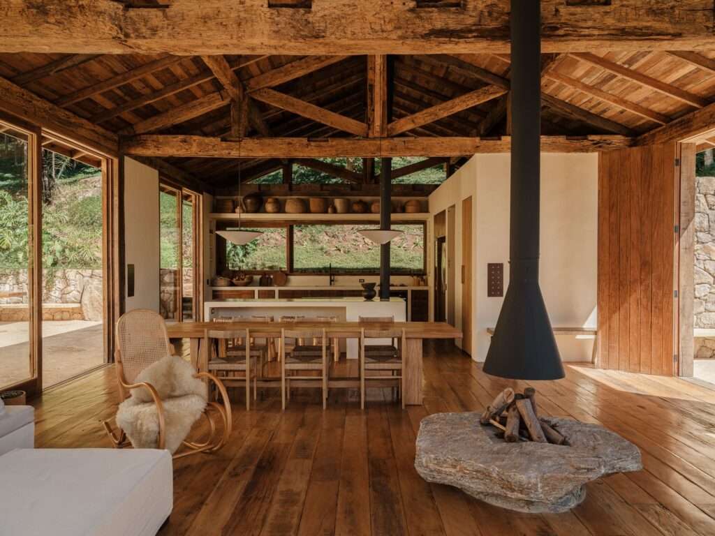Interior social living area of Japú House featuring reclaimed timber trusses and a central suspended metal fireplace with a raw stone base.