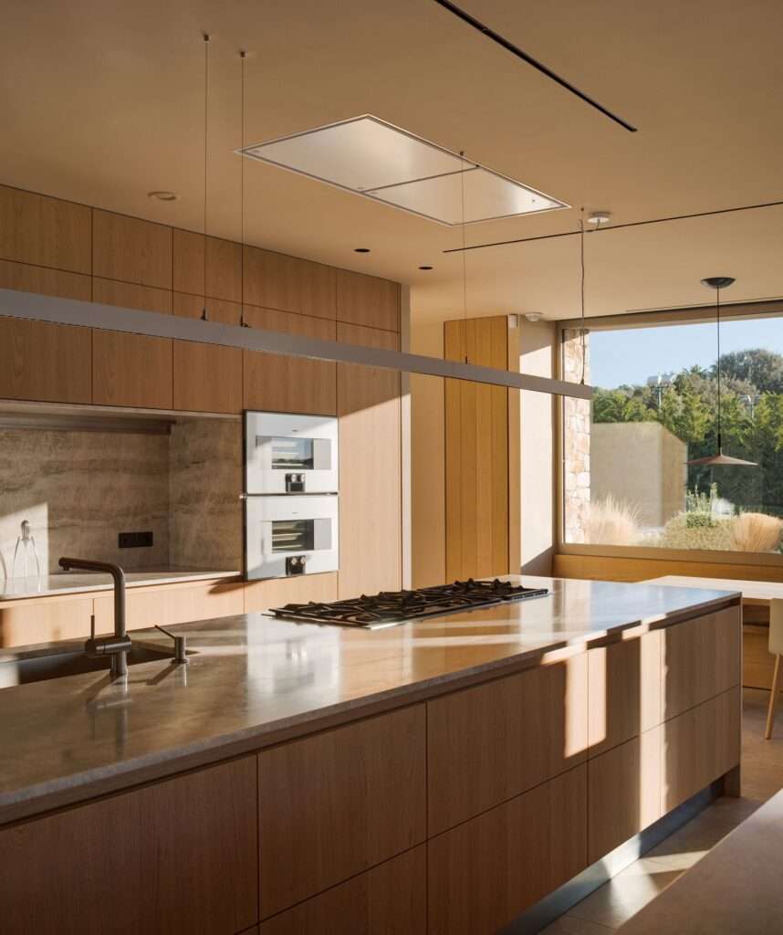 Contemporary kitchen with a massive stone-topped island, wooden cabinetry, integrated appliances, and large scenic window.