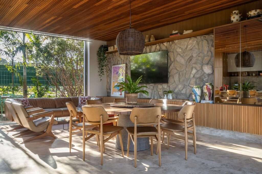 Round wooden dining table with chairs set against a natural stone wall with mounted television and decorative art.