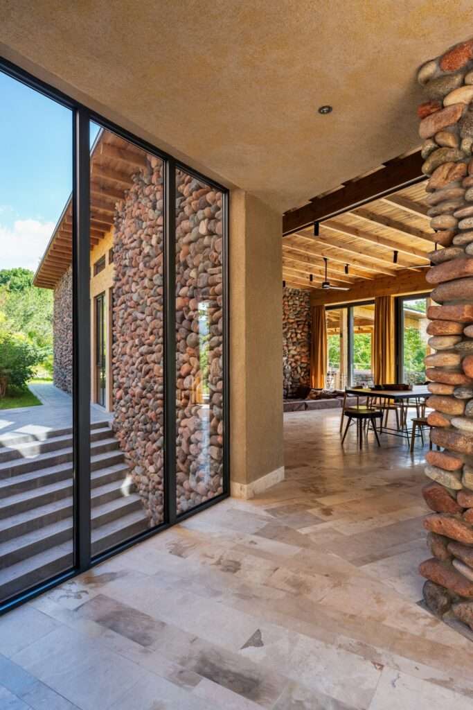 Transitional interior hallway showing stacked stone column, light stone floor, and glass wall overlooking an exterior staircase.