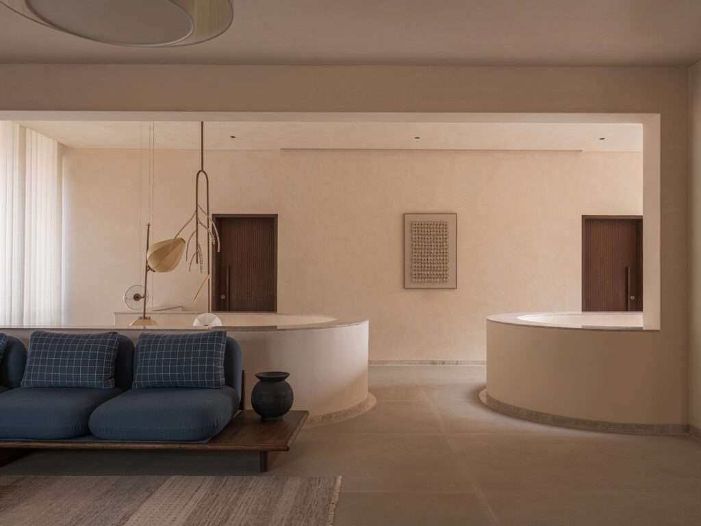 Upper-floor living room with beige plaster walls, custom blue modular sofa, and curved ceiling cutouts overlooking the central vertical light atrium.