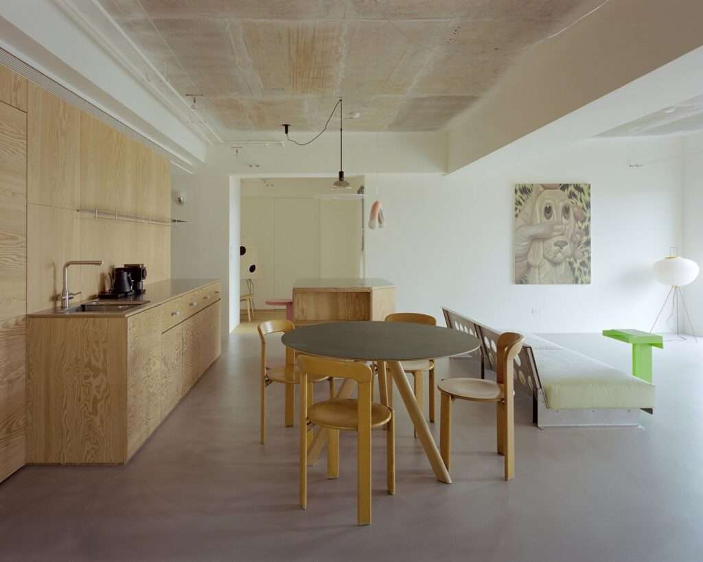 Open-plan apartment interior showing dining table, wall art of a dog, paper floor lamp, pine plywood kitchen counters, and exposed raw concrete ceiling.