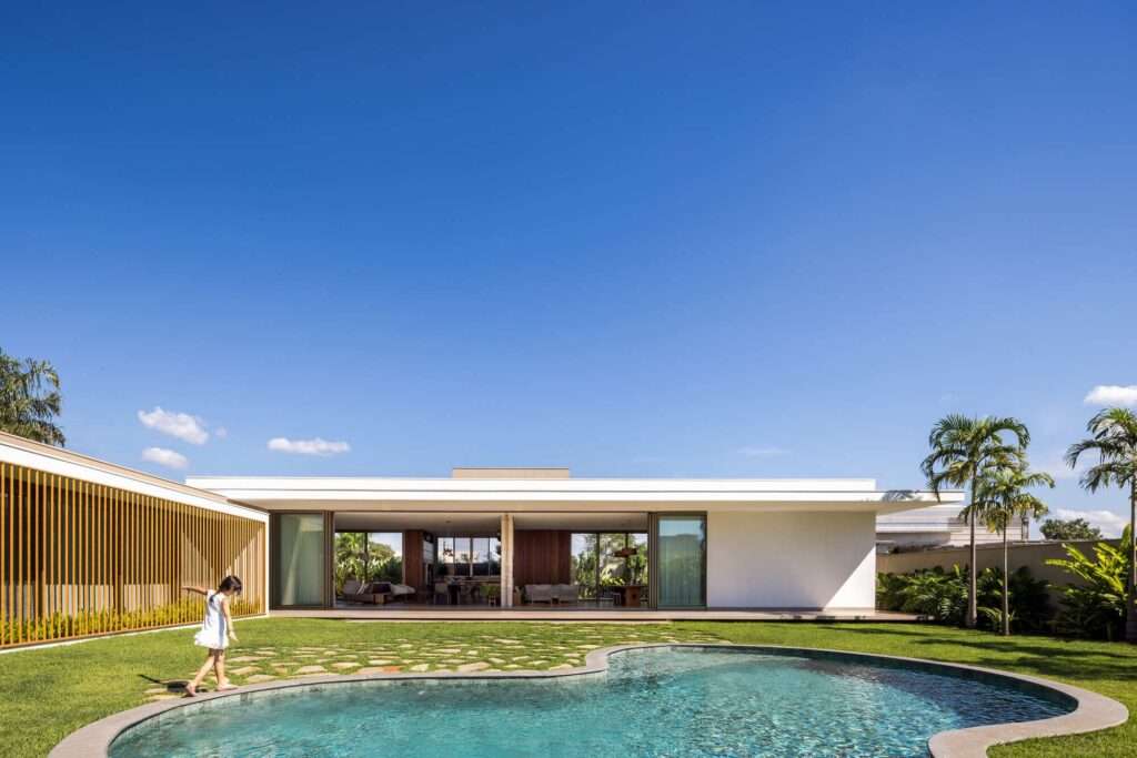 Full view of the L-shaped Refugio House facade with open living pavilion, organic curved swimming pool, lawn, and vertical wooden solar louvers.