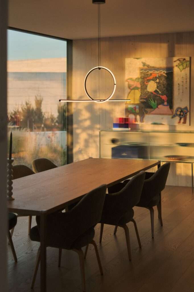 Interior view of an elegant dining room in Naseem House featuring a solid wood dining table, dark green upholstered chairs, a modern circular pendant light, and a large window overlooking a golden field at dusk.