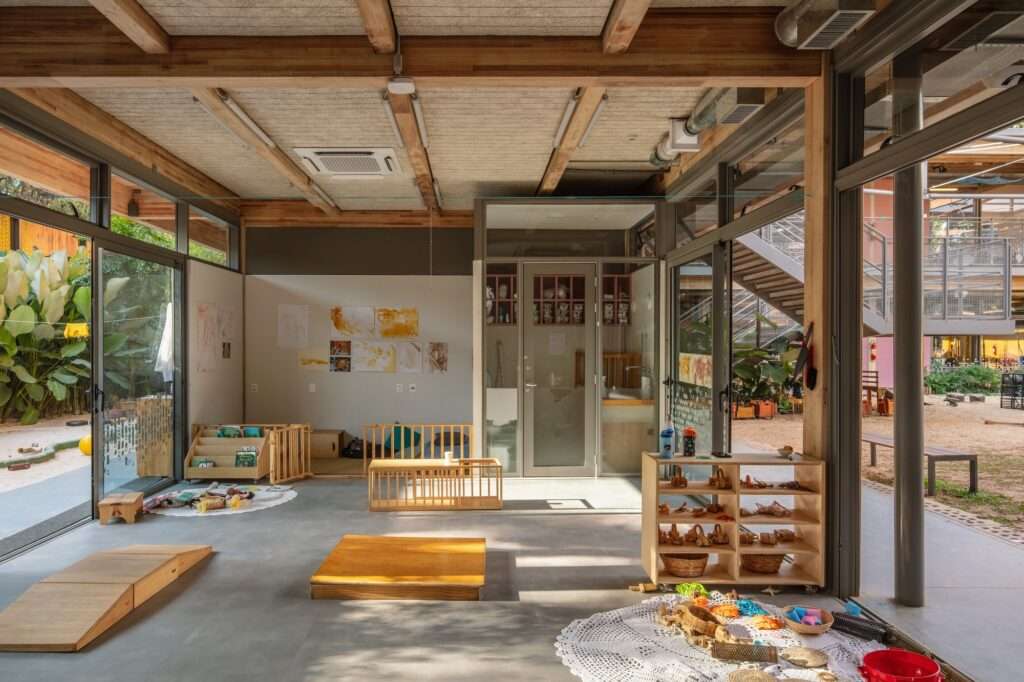 Early childhood playroom interior with modular low-height wooden shelves, play mats, exposed wooden beams, and full-height glass windows leading outdoors.