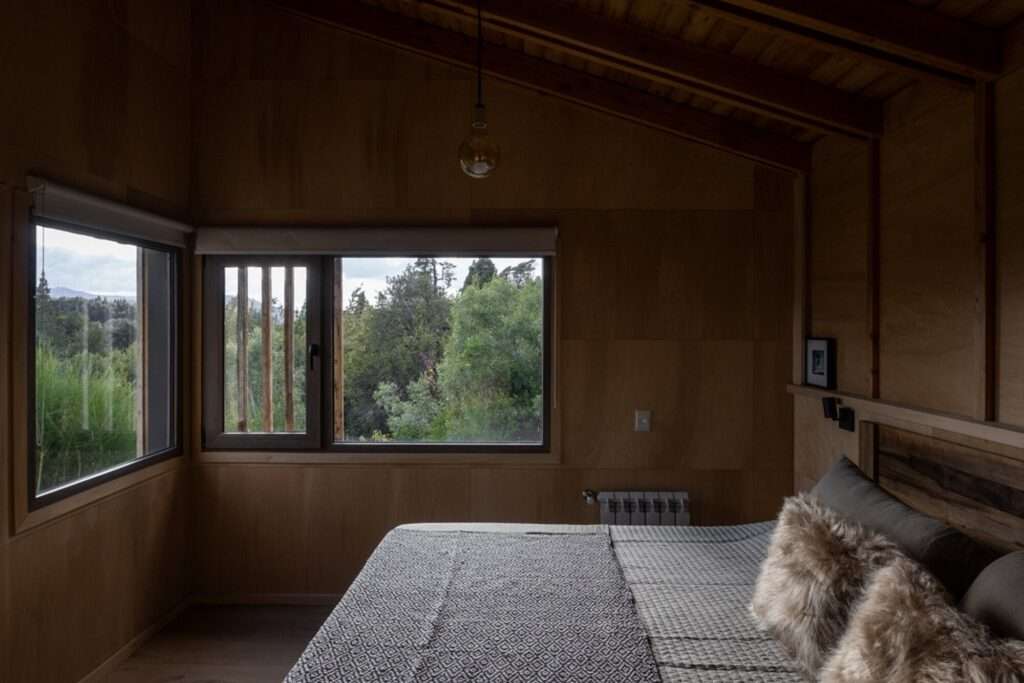 Bedroom interior featuring Guatambu timber walls, sloped wooden ceiling, and a framed window with vertical wooden louvers facing trees.
