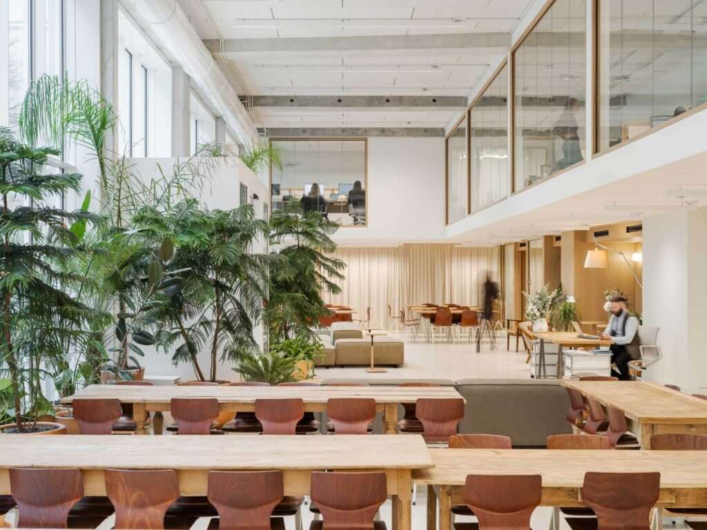 Bright workspace interior with long wooden communal tables, indoor greenery, and glass-partitioned upper floor meeting rooms.
