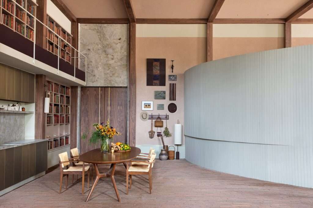 Curved light grey partition wall sweeping past a dining setup and displaying rural heritage items on an earthy pink wall.