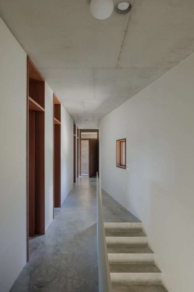 A view down a long, narrow hallway in EA House with white walls and a concrete ceiling, featuring deep timber door frames, a concrete floor, and stairs leading down.