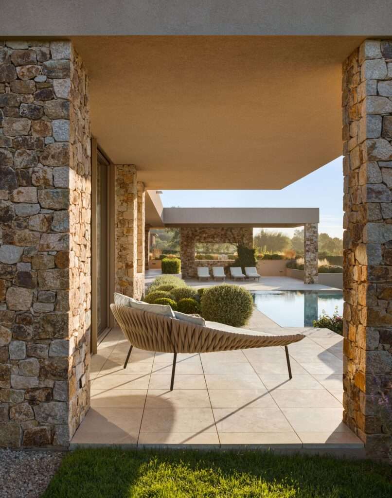 Covered stone patio with modern outdoor lounge chair overlooking a swimming pool and sun loungers.