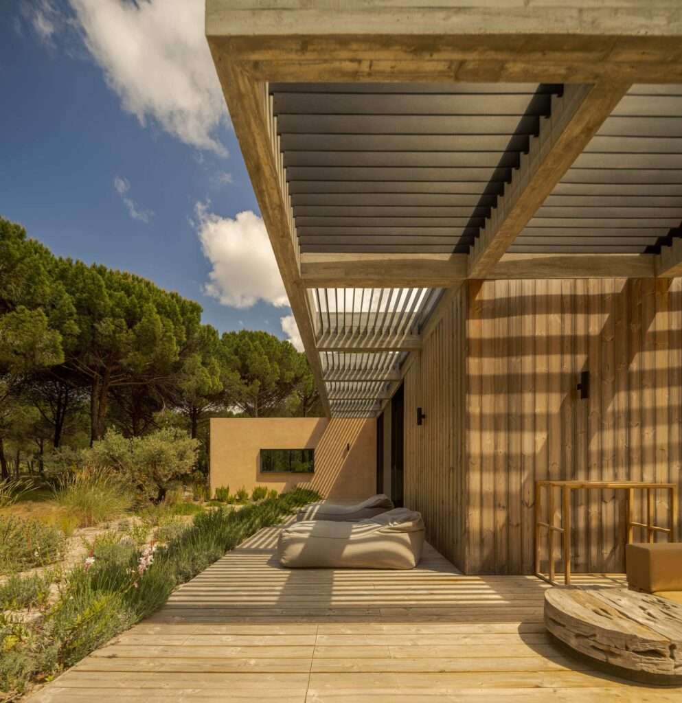 Shaded wooden terrace under concrete pergola casting linear shadows on Muda L13 wall cladding.