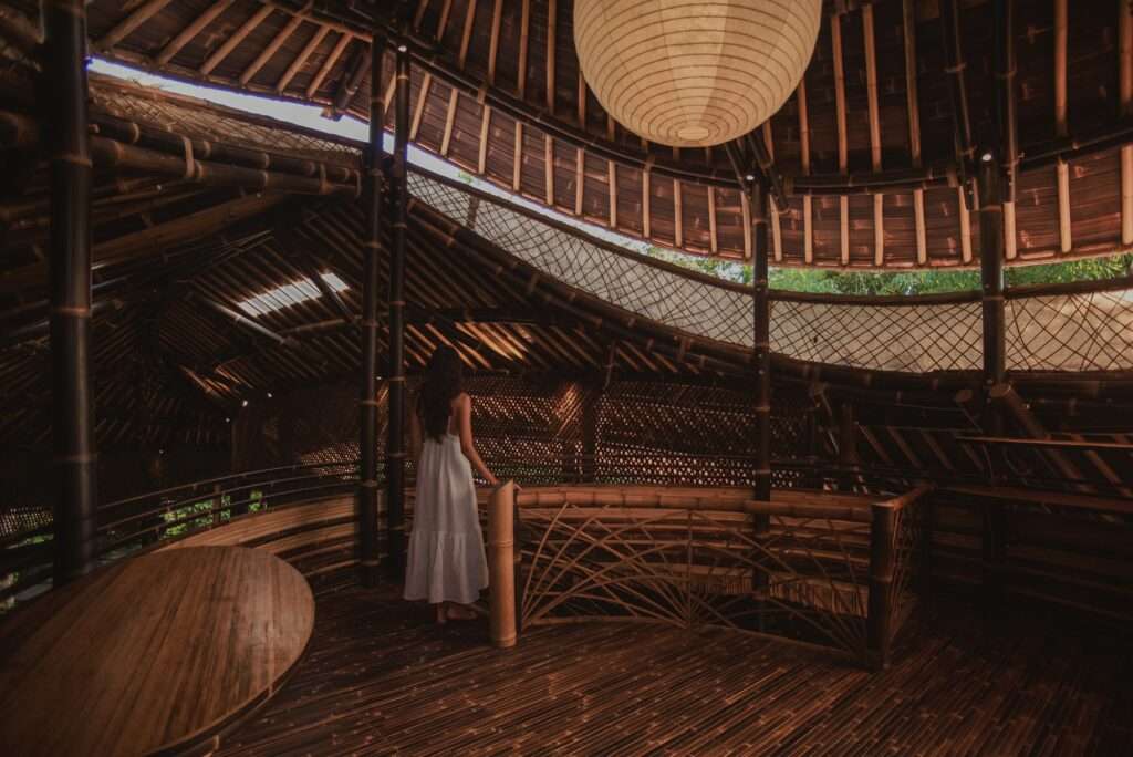 Interior view of the upper mezzanine level at night, showing a person with long dark hair looking out from a bamboo-railed balcony with string lights, within a large open-plan bamboo and tile-roofed structure.