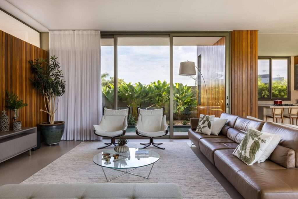 Modern interior living room featuring a leather sofa, wooden wall cladding, neutral rug, and large sliding glass doors overlooking tropical courtyard greenery.