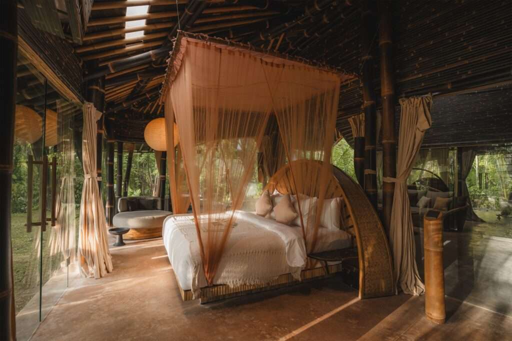 Interior view of a bedroom featuring a canopy bed with sheer curtains, set against a large curved glass wall looking out to a dense tropical forest, with a unique patterned ceiling and bamboo structure.