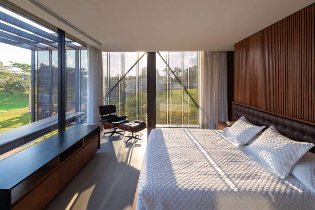 Master bedroom interior with wood wall paneling, a king-size bed, Eames lounge chair, and glass facade revealing diagonal structural tension bracing.