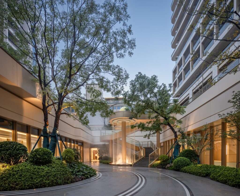 Sunken courtyard with central illuminated water feature fountain, spiral staircase, and lush green plants surrounded by residential building walls.