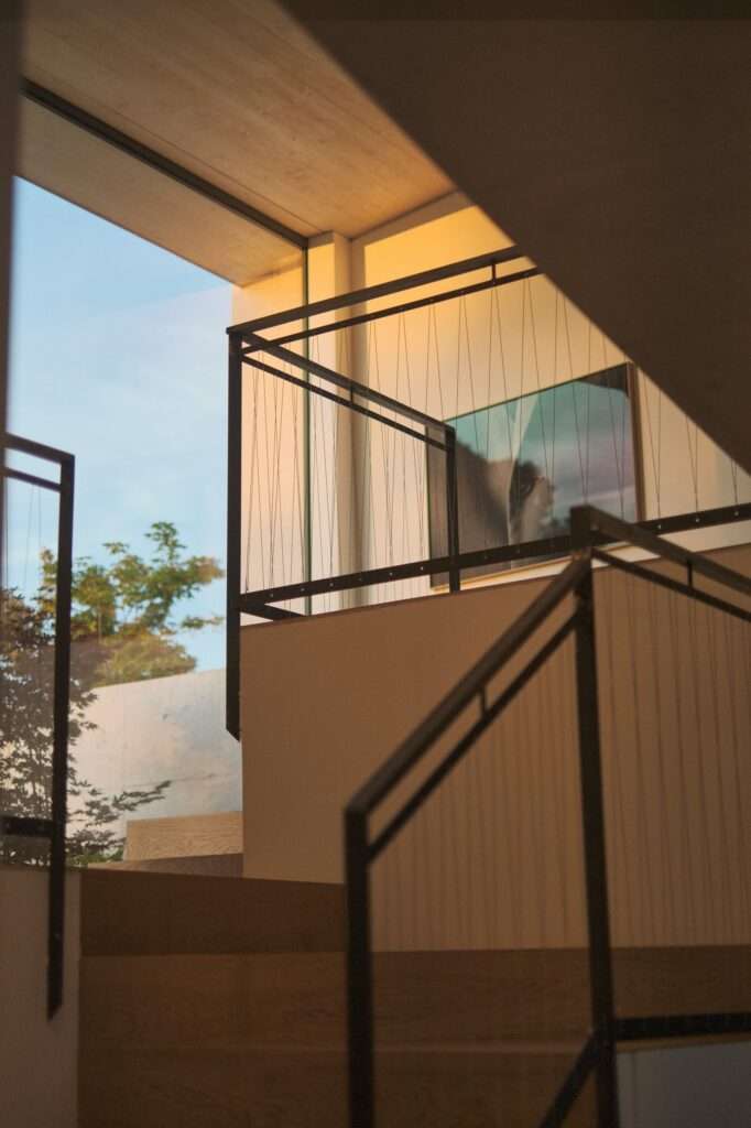 Interior detail shot looking up a wooden staircase with thin black wire cable railings, bathed in warm ambient light with a large artwork of a horse visible on the upper wall and a window framing outdoor foliage.