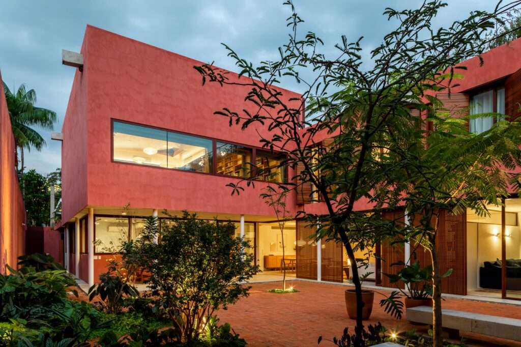 Evening dusk view of the illuminated L-shaped house courtyard showing warm interior lighting, lush garden trees, and wooden sunshades.