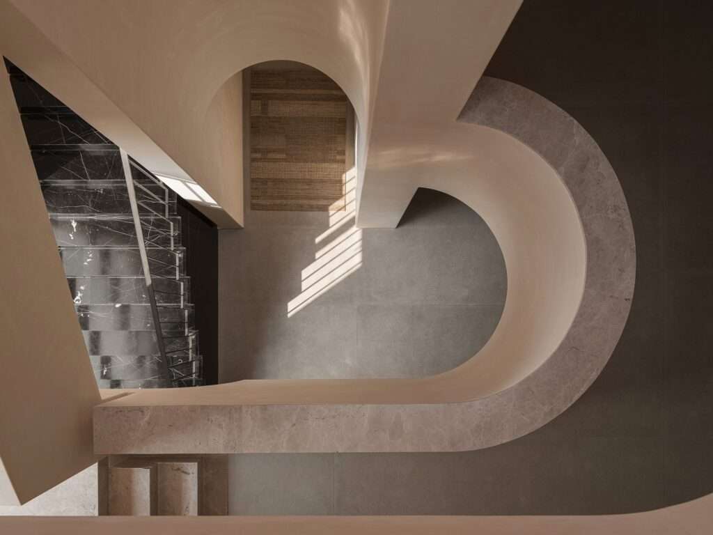 Looking down a central vertical atrium void with a sculptural curved staircase railing and dark marble steps lit by soft overhead natural light.