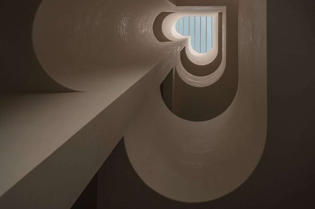 Dramatic vertical upward view through a multi-story atrium void with organic curved plaster openings terminating at a slatted glass skylight.
