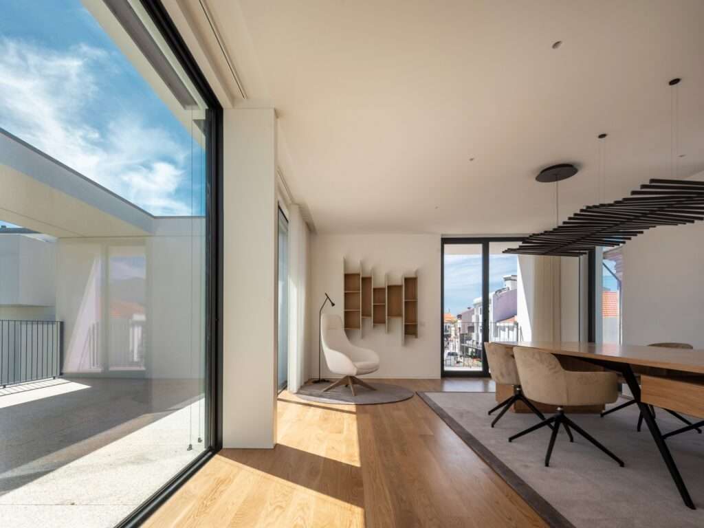 Bright modern living room interior with warm wood flooring, large glass sliding doors, and open views to an outdoor terrace and city street.