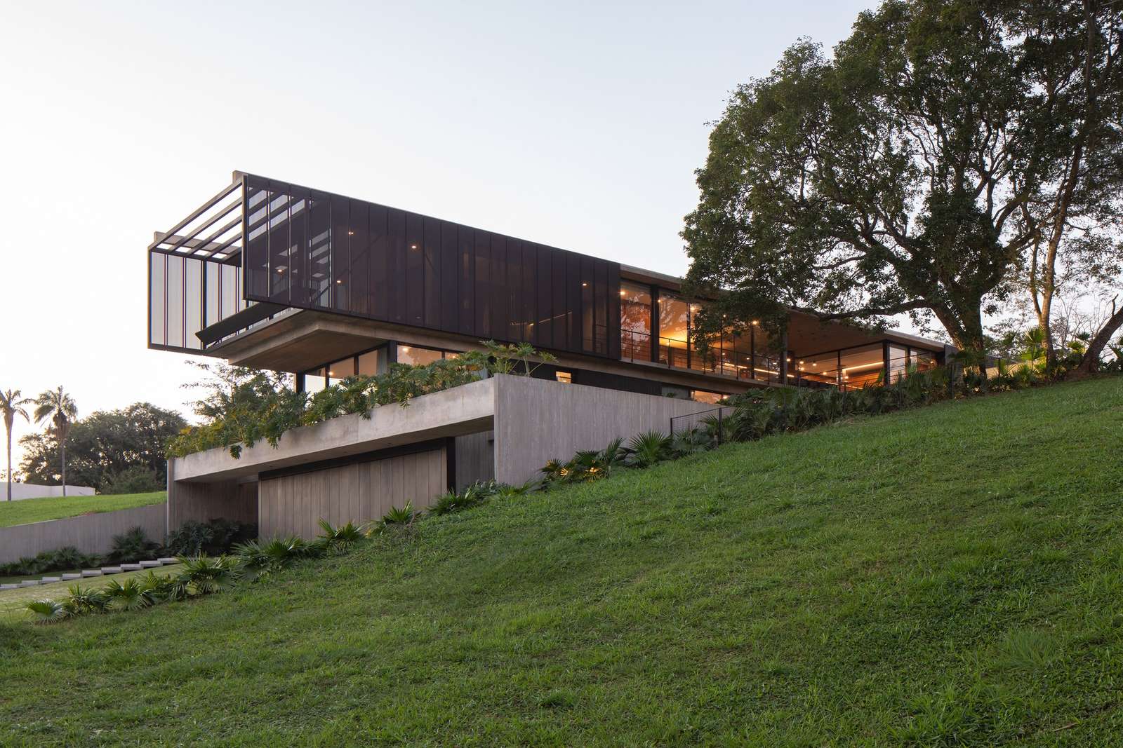 Wide exterior view of Casa Galería built into a grassy green slope, showing the tiered concrete foundation and cantilevered upper floor at dusk.