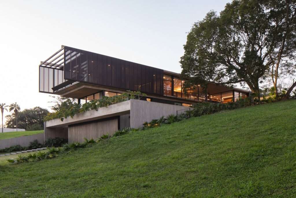 Exterior profile of Casa Galería illuminated at sunset on a grassy hill, showcasing the glow from the glazed social level beneath a large shade tree.
