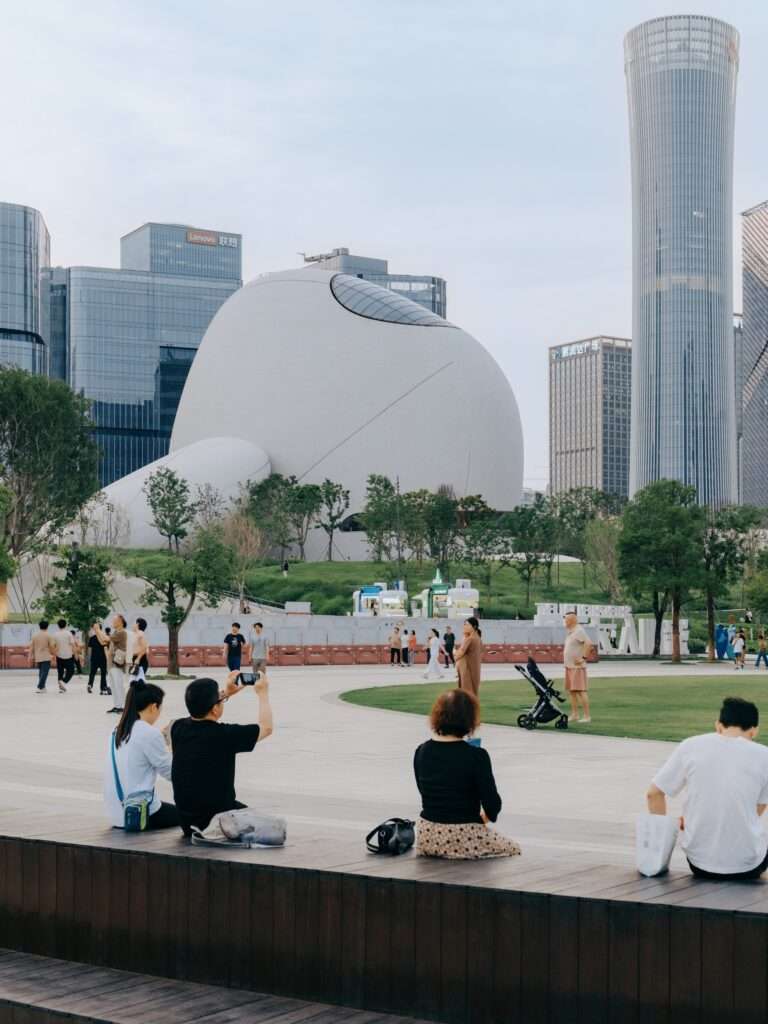 People relaxing on outdoor wooden bench seating in an open public plaza with Shenzhen Bay Culture Square pavilions and skyscrapers in the background.