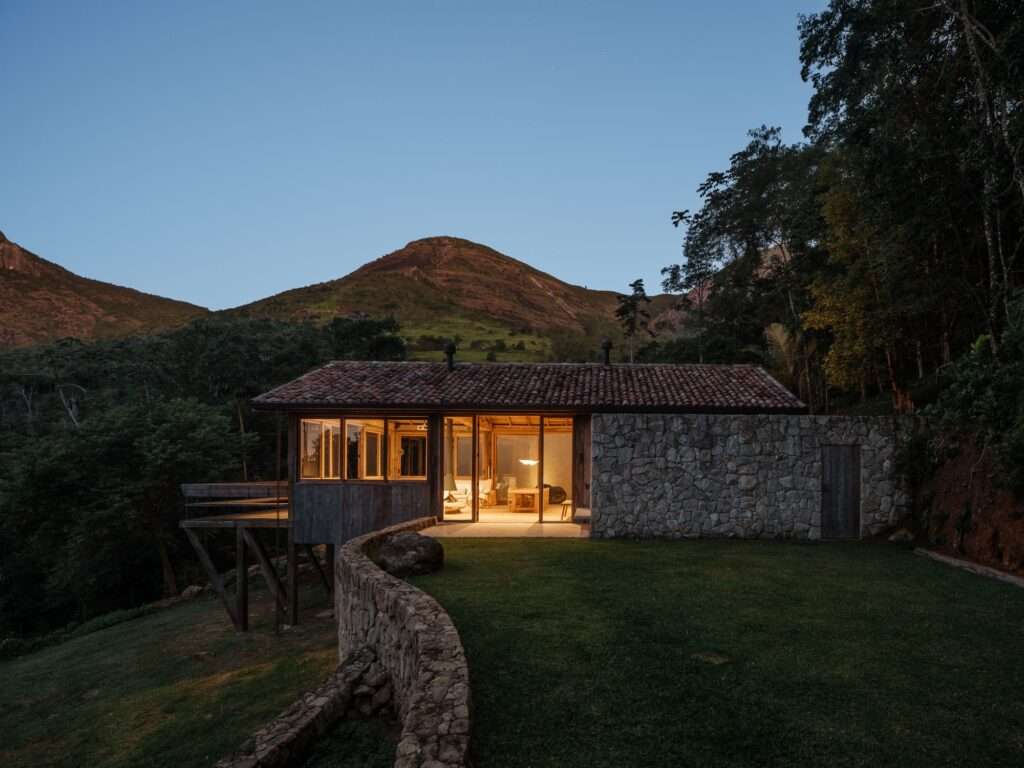 Exterior dusk photo of Japú House social pavilion illuminated against mountain backdrop with local stone retaining wall.