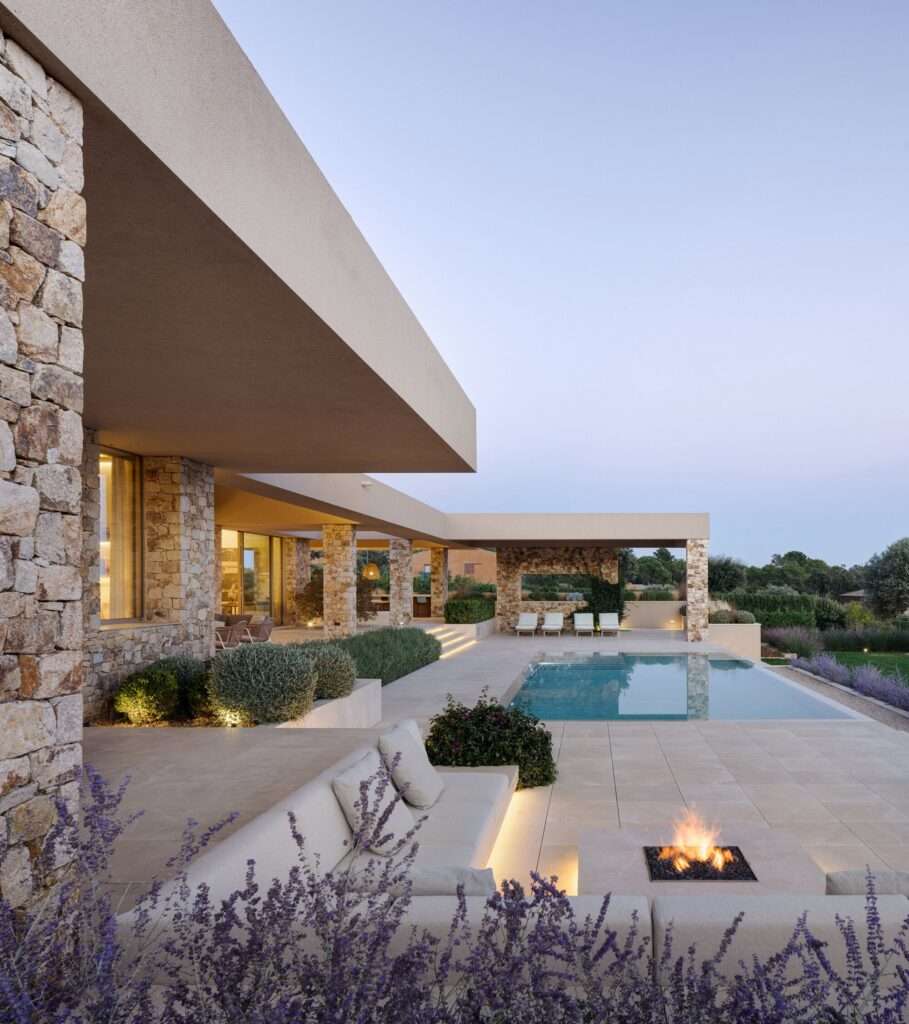 Dusk view of Talleru House outdoor patio featuring an integrated fire pit, built-in sofa, lit pool, and lavender garden.