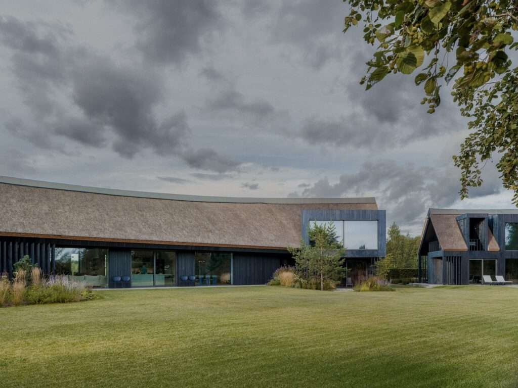 Wide panoramic view of the curved BPROM house with long thatched roof, expansive green lawn, and dark blue timber facade under cloudy sky.