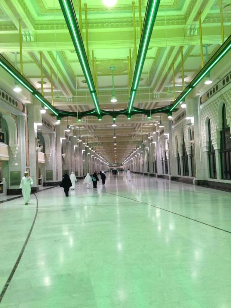 Interior view of the expanded corridors in Al-Masjid Al-Haram, featuring polished marble floors, arched columns, and modern green linear ceiling lighting.