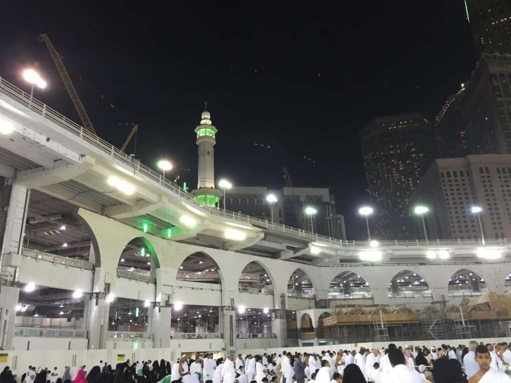 Night view of the temporary elevated circular Mataf structure encircling the Kaaba in Mecca, featuring illuminated arches and a green minaret against a dark sky.