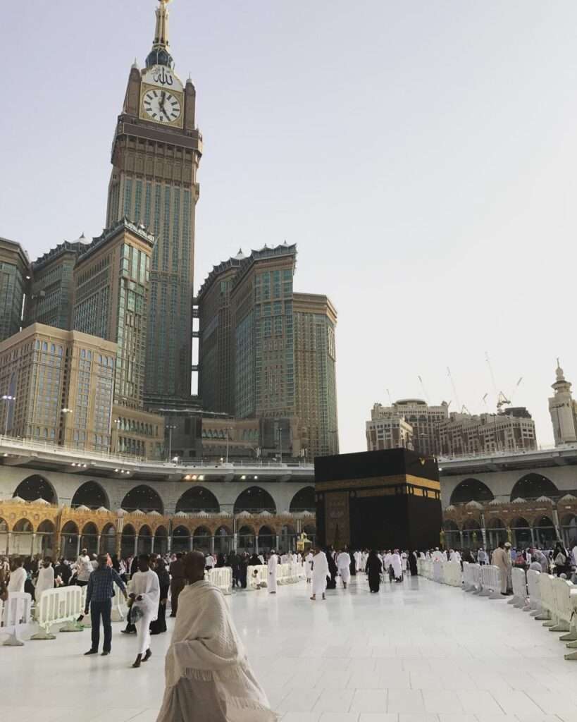 The holy Kaaba surrounded by pilgrims performing Tawaf during the day, enclosed by a temporary elevated walkway and the towering Abraj Al Bait clock tower in the background.