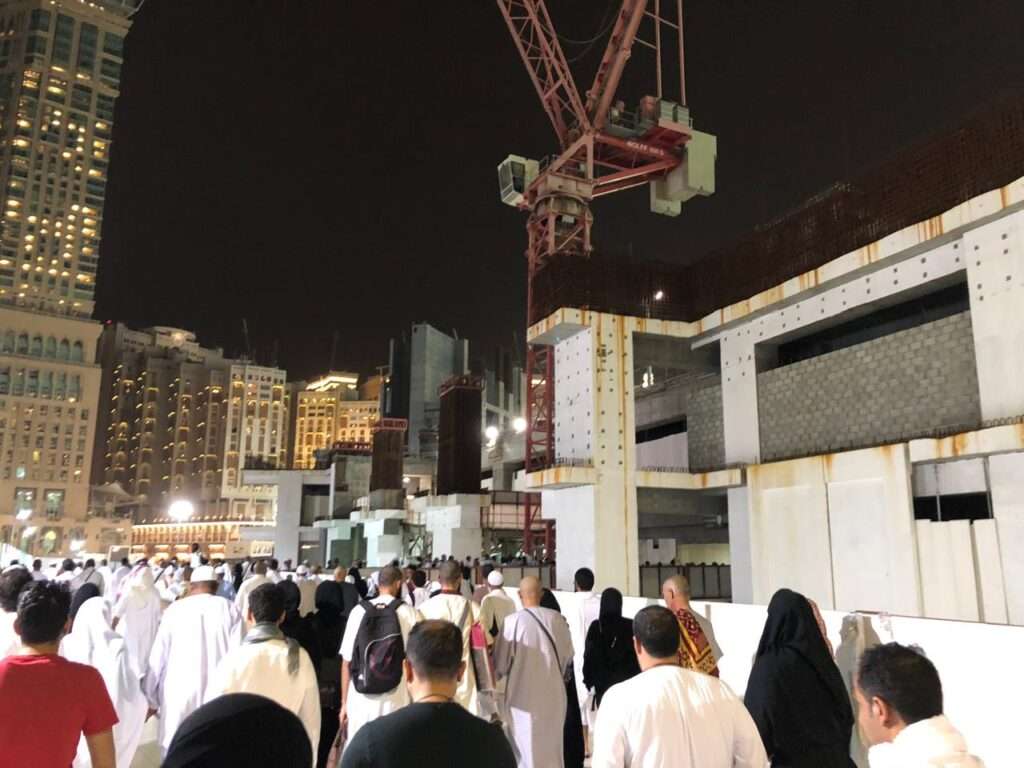 Thousands of pilgrims gathered around the Kaaba at night, with multiple massive construction cranes operating in the background over partially built mosque structures.
