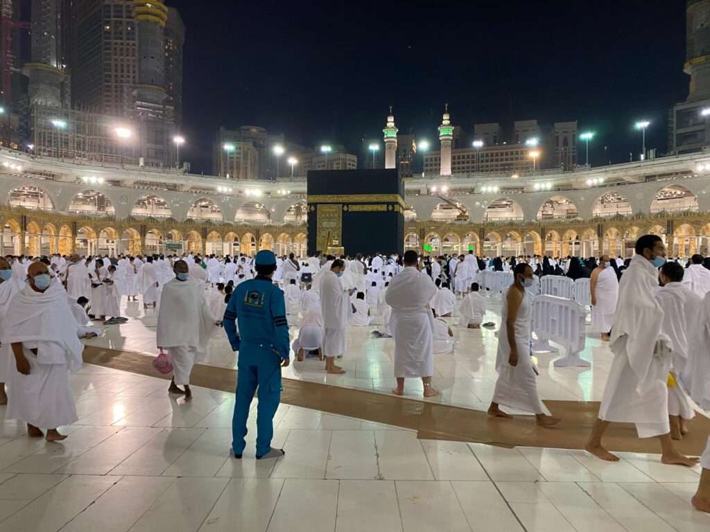 Pilgrims walking at night past a massive construction zone with exposed concrete pillars, steel rebar, and a towering red crane inside the Grand Mosque complex.