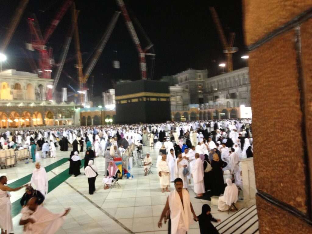 Pilgrims performing Tawaf around the Kaaba at night on polished white marble, with traditional illuminated archways and minarets in the background.