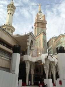 Daytime view of the partial demolition and reconstruction of the Grand Mosque's historical porticos, showing exposed rebar on white pillars against the backdrop of the Makkah Clock Tower.