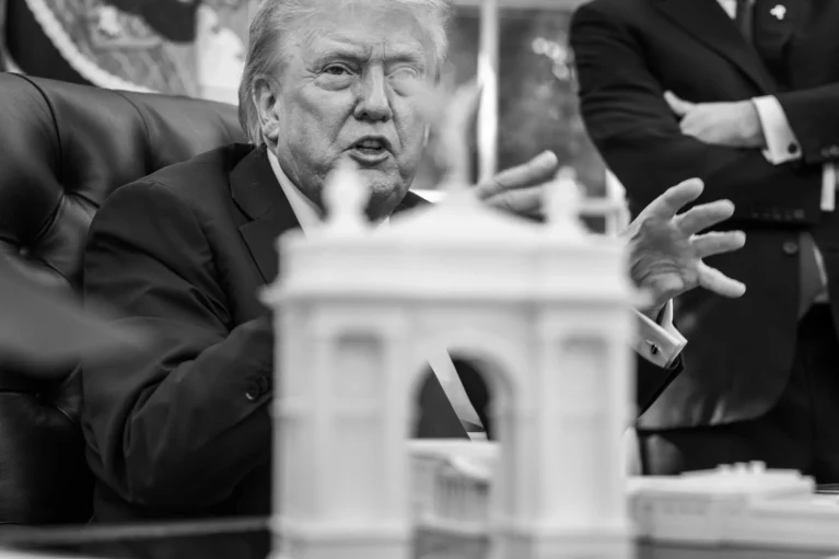 Donald Trump gesturing seated behind a white physical architectural model of a classic triumphal arch structure
