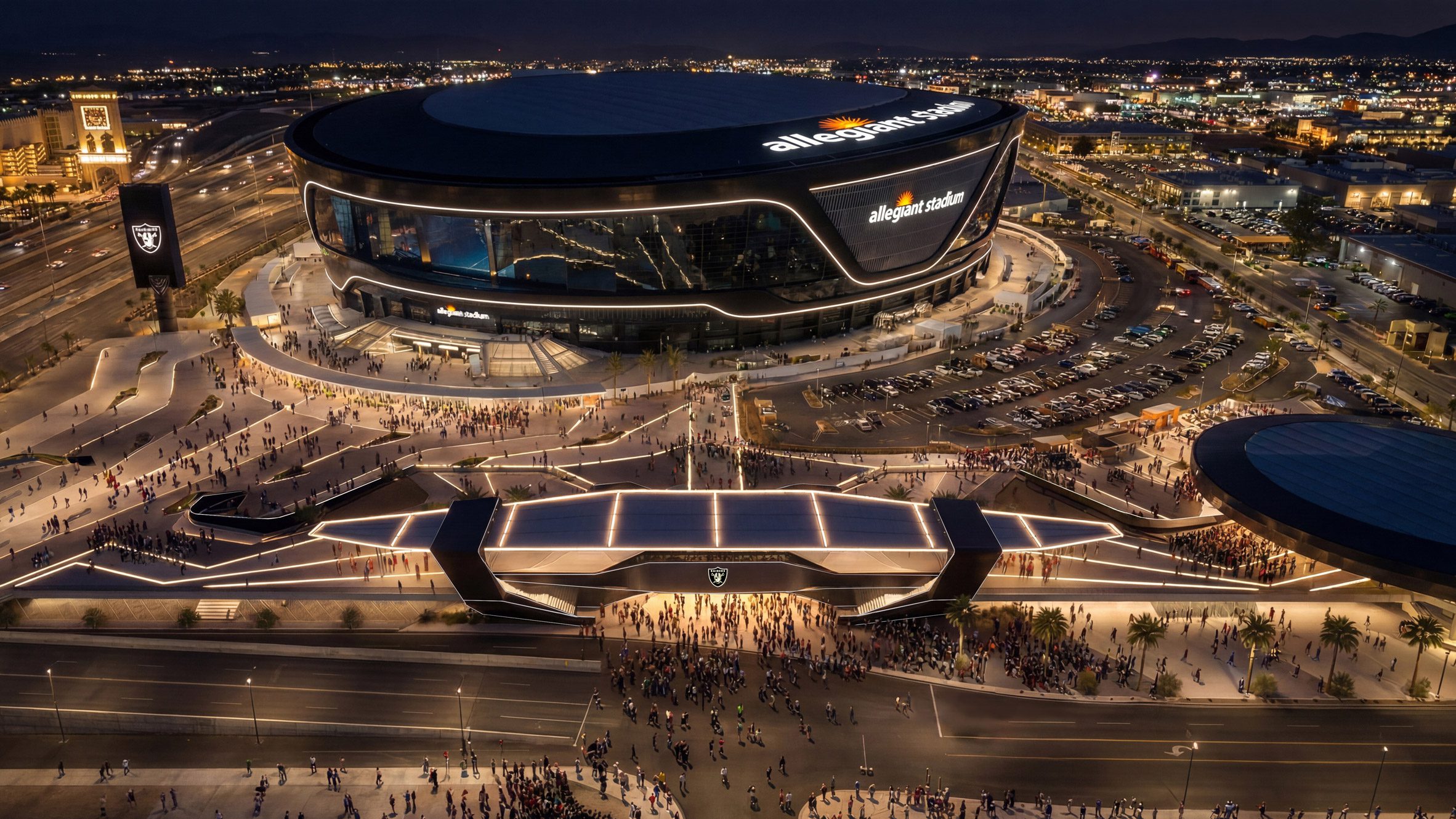 Night elevated view showing Allegiant Stadium alongside illuminated multi-level entry plaza infrastructure and surrounding arterial roadways.