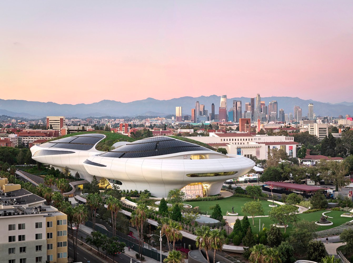 Aerial view of the curved Lucas Museum building surrounded by green parkland and the Los Angeles skyline.