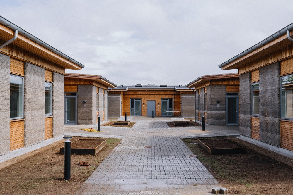 Paved circulation route framed by single-story 3D-printed concrete and timber residential buildings.