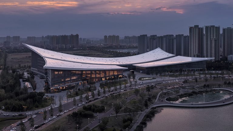 Elevated view of a modern library complex with a sweeping curved white roof illuminated at dusk.