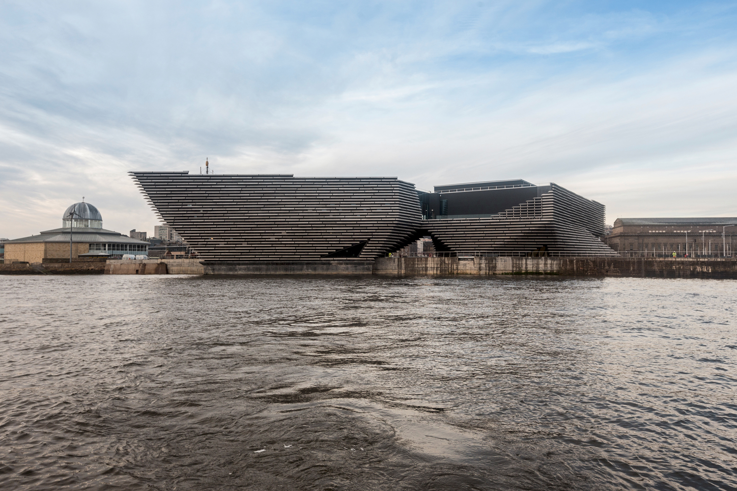 Exterior view of the V&A Dundee building featuring horizontal stone cladding modules over water.