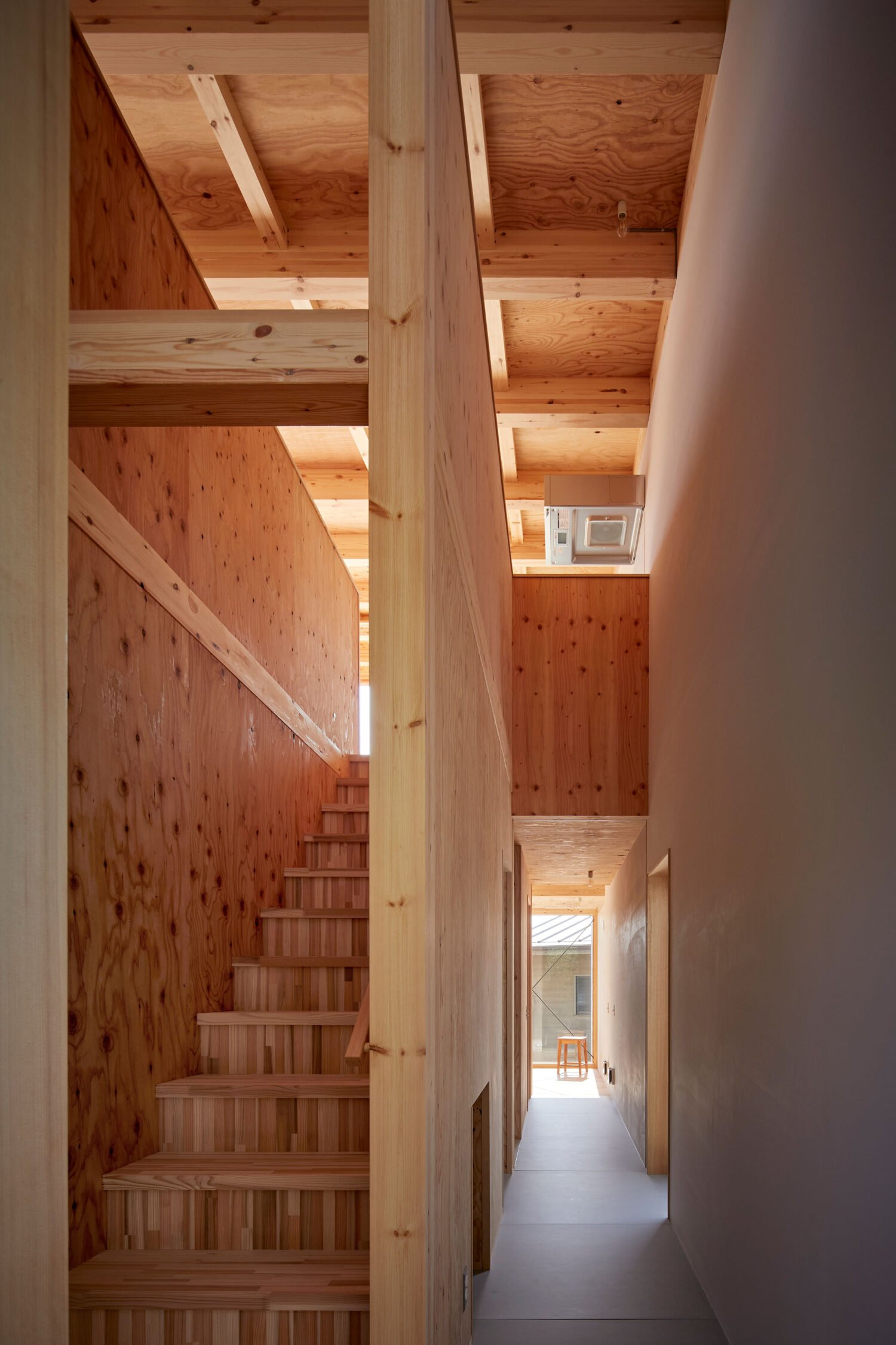 Narrow interior corridor with exposed timber ceiling joists, wooden stairs, and plywood wall finishes.