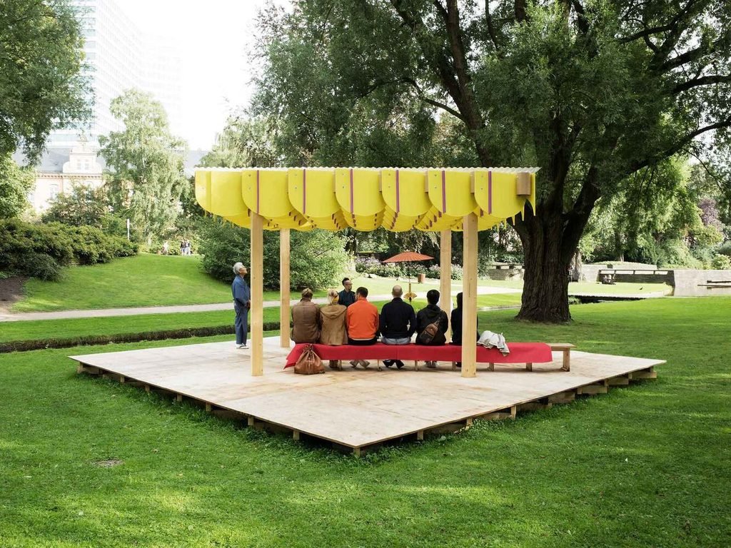 People seated on benches under a wooden pavilion canopy on a square timber platform.