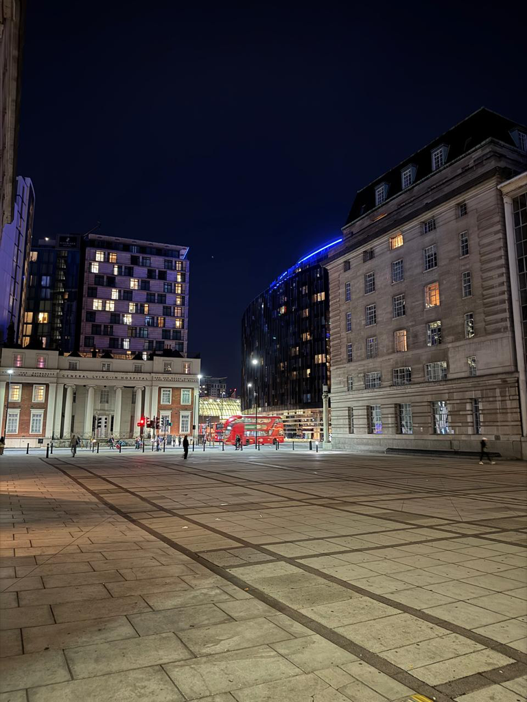 A wide, grid-paved urban plaza at night, leading to a street with a moving red double-decker bus. The square is flanked by the classical, columned facade of the historic General Lying-In Hospital on the left and a large, monumental stone building on the right, contrasted against modern illuminated high-rises.